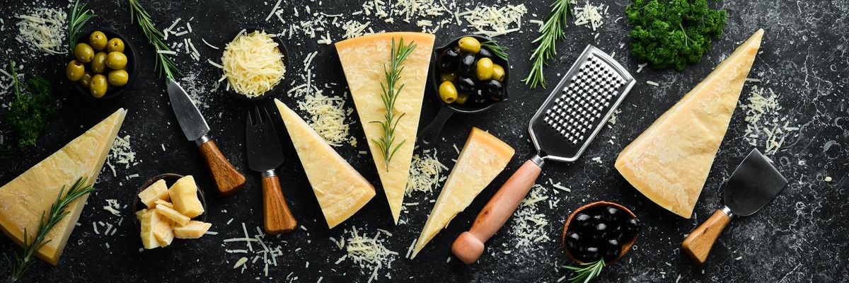 cheddar sajtok fekete asztallapon, körülöttük reszelő, kés, olivabogyók