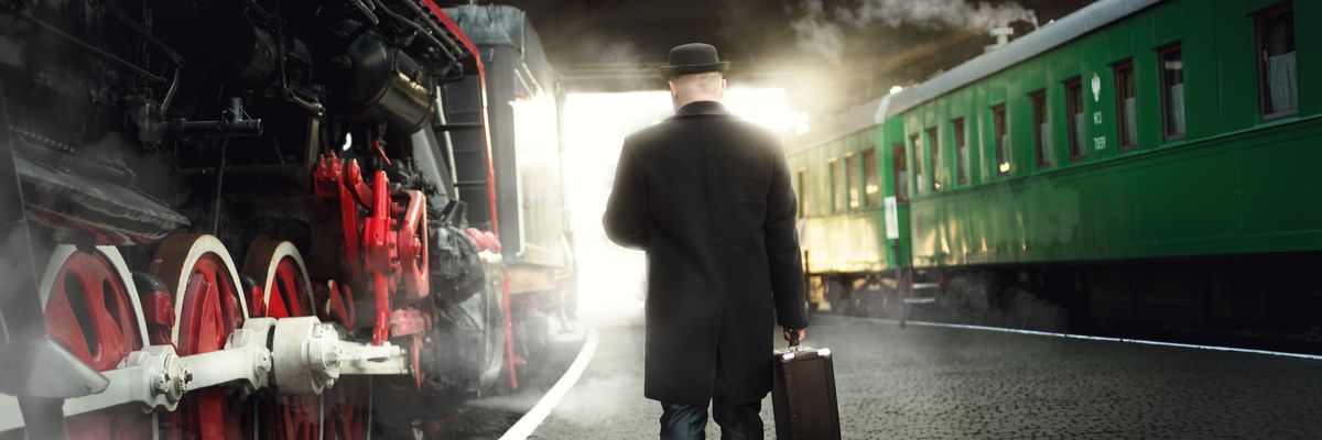 egy fekete kabátos, kalapos férfi sétál bőrönddel egy régi gőzmozdony mellett