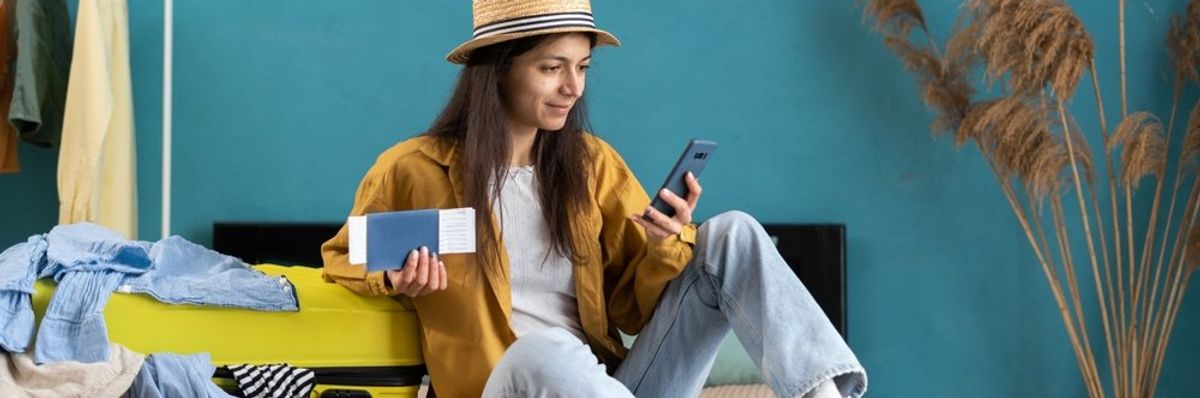 egy fiatal nő a bepakolt bőröndje mellett ül kalapban, repjeggyel a kezében, és a telefonját nézi