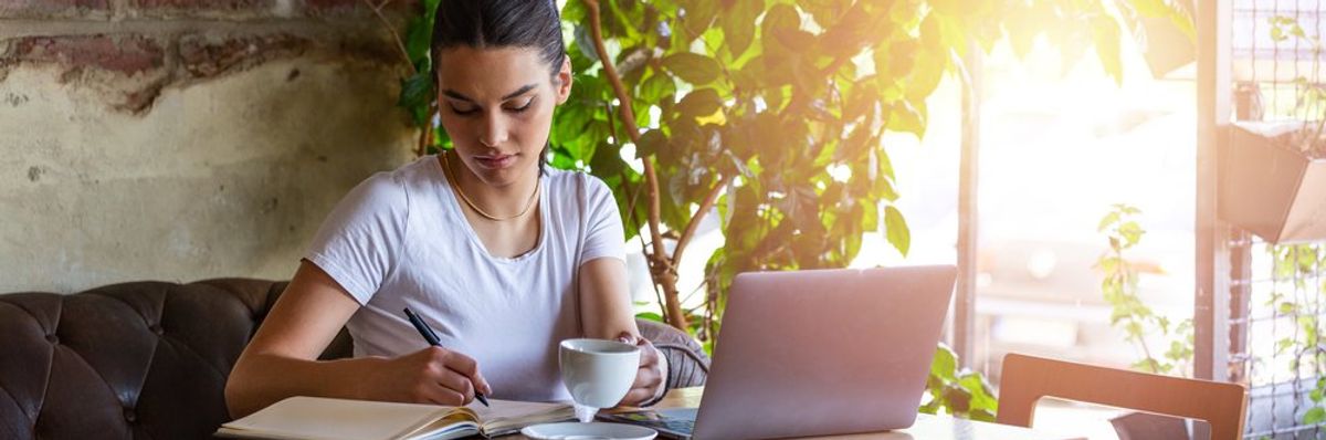 egy nő dolgozik egy kávézóban, előtte a laptopja, ír egy noteszba, fél kézzel pedig felemeli a kávéscsészéjét