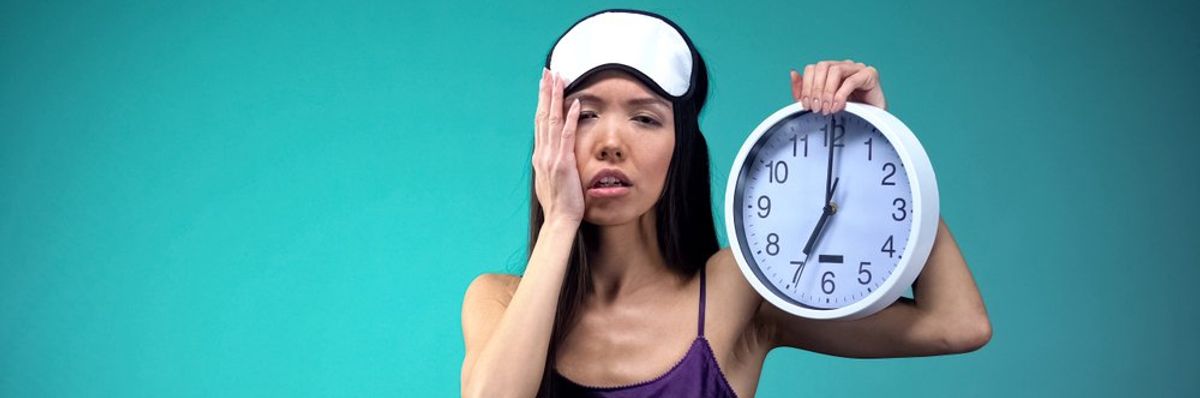 egy nő egy 7 órát mutató faliórát fog a kezében álmosan, fején alvómaszk