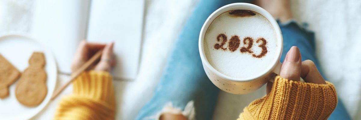 egy nő fog egy csészét, amiben kávéhabon 2023 felirat van kakóporral írva, mellette egy füzetbe ír