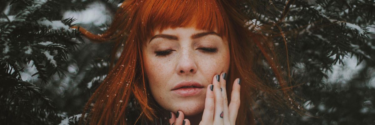 egy vörös hajú nő lehunyt szemmel at arcához érinti az ujjait a fenyőfák előtt, a haján hópelyhek