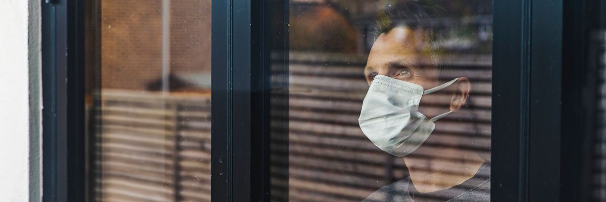 férfi szürke pulóverben és az arcát takaró maszkban egy ablakban áll és kifelé mered a reluxa mögül