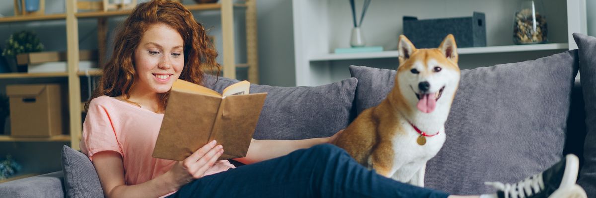fiatal lány a kanapén fekve mosolyogva könyvet olvas, mellette a kutyáját simogatja