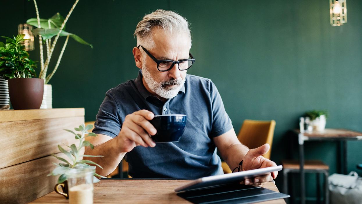 idős ősz szakállas férfi kávét a kezében tartva olvas valamit egy tableten vagy ipaden egy kávézó fa asztalánál ülve növényekkel körülötte