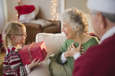 szőke szemüveges kislány kötött pulóverben piros csomagolású aranyszalagos ajándékot nyújt át idős nagymamájának aki zöld felsőt visel mellete ül a kanapén a piros pulóveres papa