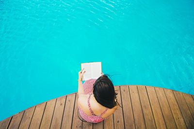 Fiatal nő egy medence partján ül a stégen, és egy könyvet olvas