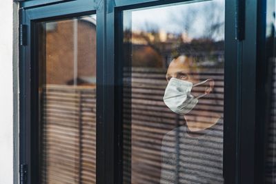 férfi szürke pulóverben és az arcát takaró maszkban egy ablakban áll és kifelé mered a reluxa mögül