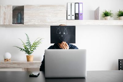 ​férfi laptop mögött mosolygó arcot ábrázoló képet tart az arca elé a kezében egy nagy szürke asztalnál rajta a telefonjával háttérben növénnyel és polcokkal a fehér fal előtt