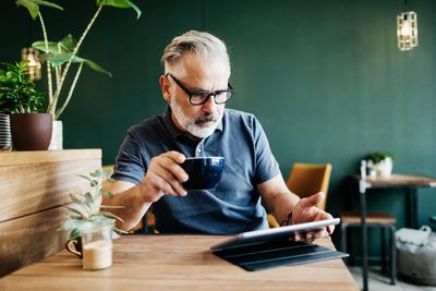 ősz szakállas férfi szemüvegben tabletet ipadet olvas kávéval a kezében egy kávézóban növényekkel körülötte egy asztalnál