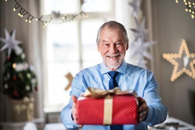 egy kék inget és sötétkék nyakkendőt viselő idős férfi egy piros csomagolású arany masnival átkötött karácsonyi ajándékot tart a kezében egy nappali szobában a háttérben egy karácsonyfával