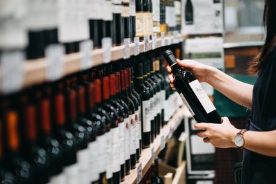 nő karórával a csuklóján egy boros polc felé nyúl és egy üveg vörösbort tart a kezében egy üzletben