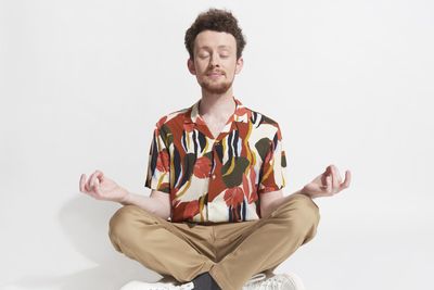 meditáló férfi mintás ingben barnás drapp színű vászonnadrágban szakállas jóképű göndör barna hajú mosolyog csukott szemmel fehér háttér előtt