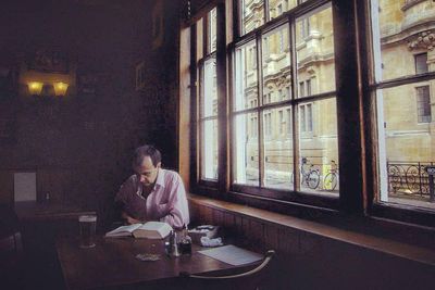 kopaszodó férfi az oxfordi Lamb and Flag nevű pubban sört iszik és könyvet olvas egy hatalmas ablak előtt asztalnál fa széken ülve rózsaszín ingben