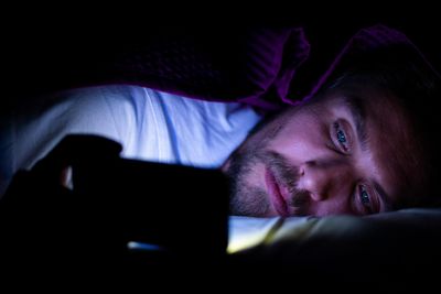 szexi jóképű szőkés szakállas kékszemű fehér pólós férfi telefonozik egy takaró lila pokróc alatt az ágyban a képébe világít az okostelefon kijelző fénye
