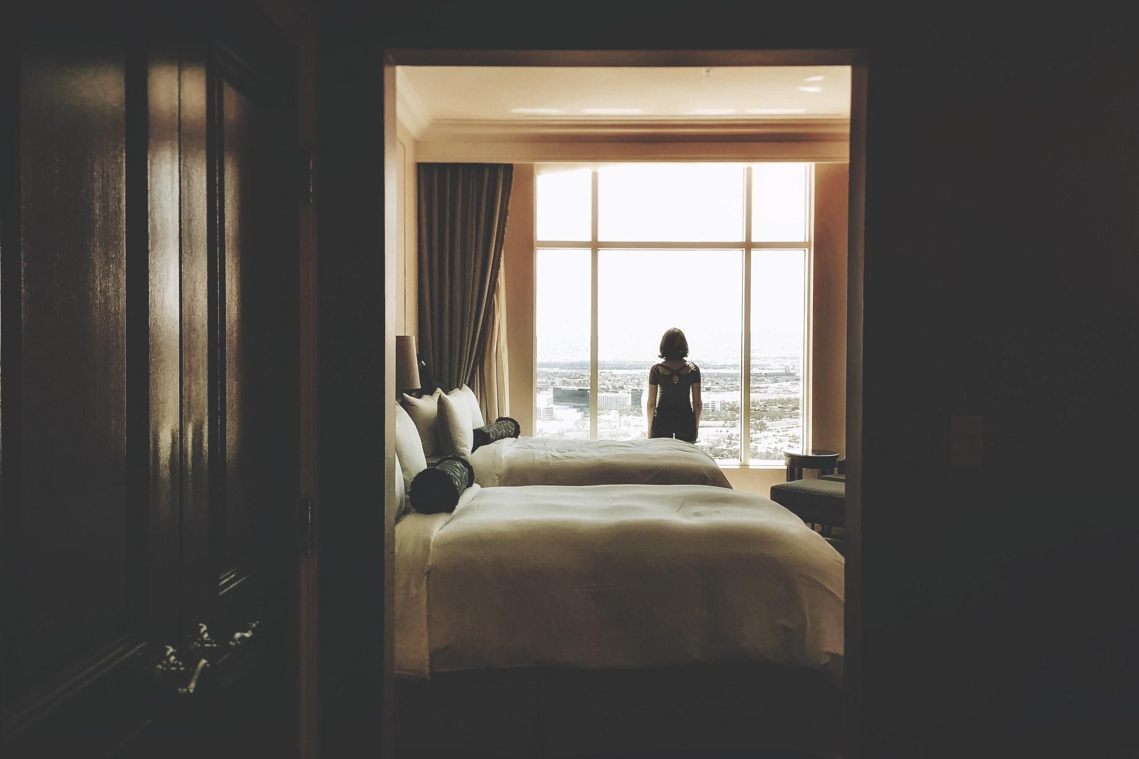 Egy nő áll egy szállodai szoba ablaka előtt háttal a kamerának