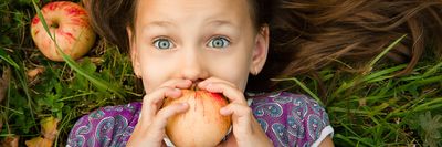 kislány almát tart a szája elé és tágra nyílt szemmel néz a fűben