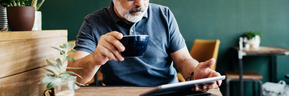 ősz hajú és szakállú szemüveges férfi kávét iszik és tabletet ipadet olvas egy kávózóban egy asztalnál körülötte növények