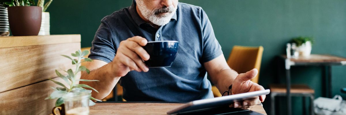 ősz hajú és szakállú szemüveges férfi kávét iszik és tabletet ipadet olvas egy kávózóban egy asztalnál körülötte növények