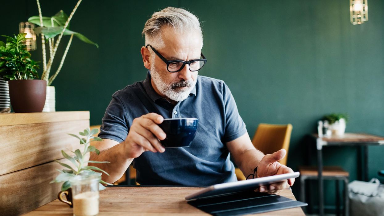 ősz szakállas férfi szemüvegben tabletet ipadet olvas kávéval a kezében egy kávézóban növényekkel körülötte egy asztalnál
