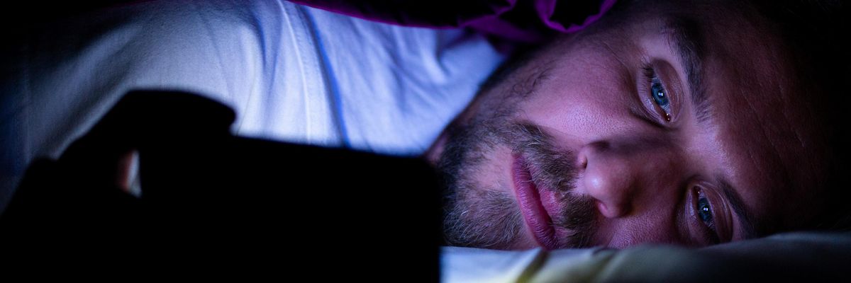 szexi jóképű szőkés szakállas kékszemű fehér pólós férfi telefonozik egy takaró lila pokróc alatt az ágyban a képébe világít az okostelefon kijelző fénye